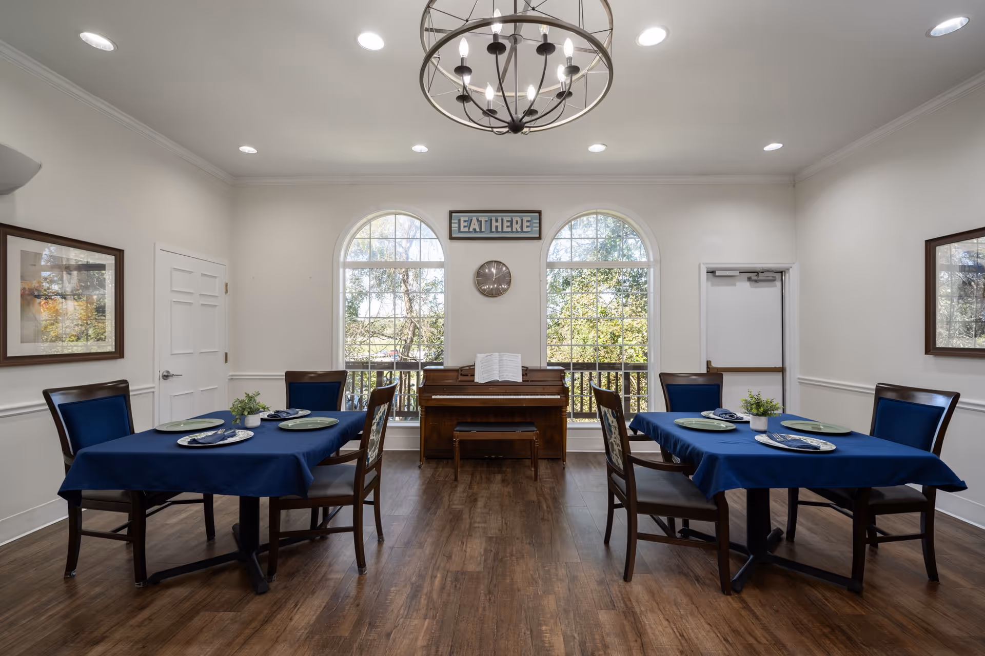A dining room with two tables covered in blue tablecloths, each set with plates and napkins. Four chairs with blue cushions surround each table. In the background, there are two large arched windows letting in natural light, a piano with an open music book, a clock on the wall, and a sign above the piano that reads 'EAT HERE'. The room has wooden flooring, white walls, and a modern chandelier hanging from the ceiling.