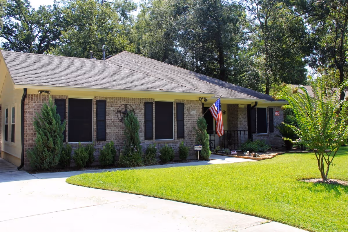 Single-story brick cottage with a front porch, American flag, shrubs, and a manicured lawn.