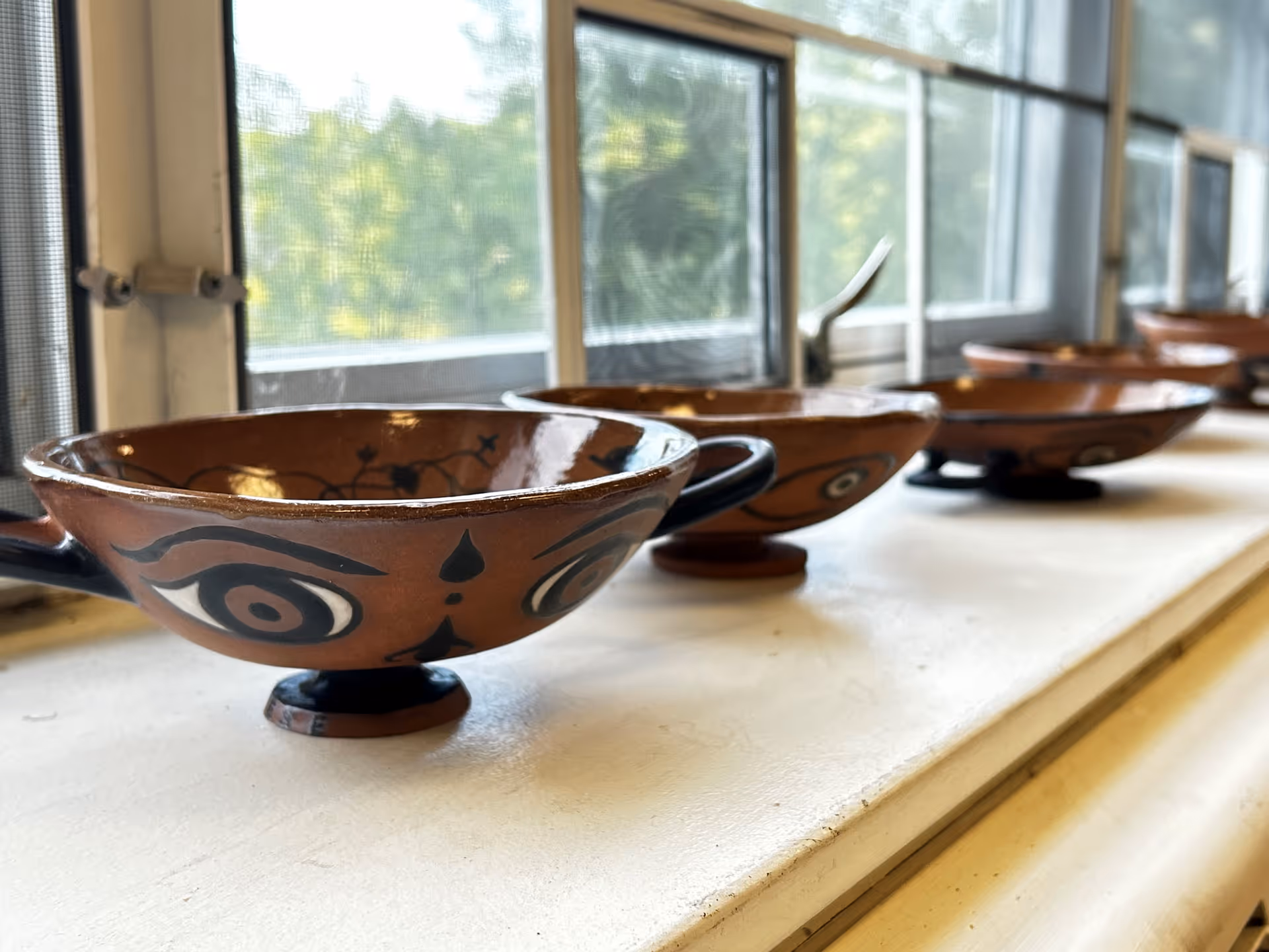 A row of decorative brown ceramic bowls with black painted designs resembling eyes, placed on a white windowsill with a window and green trees visible outside.