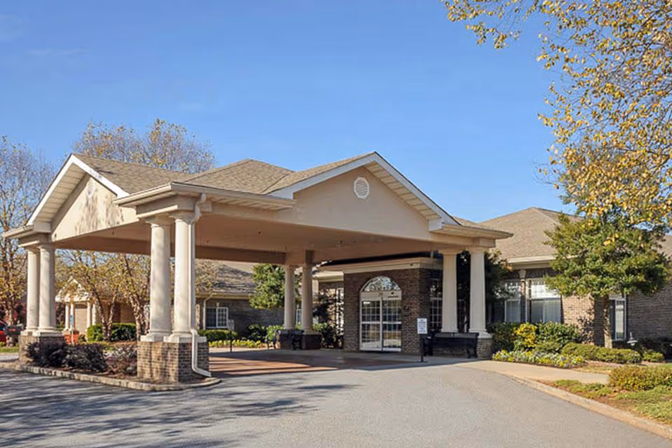 Covered porte-cochère entrance of a single-story brick senior living facility with columns, landscaping, and a driveway.