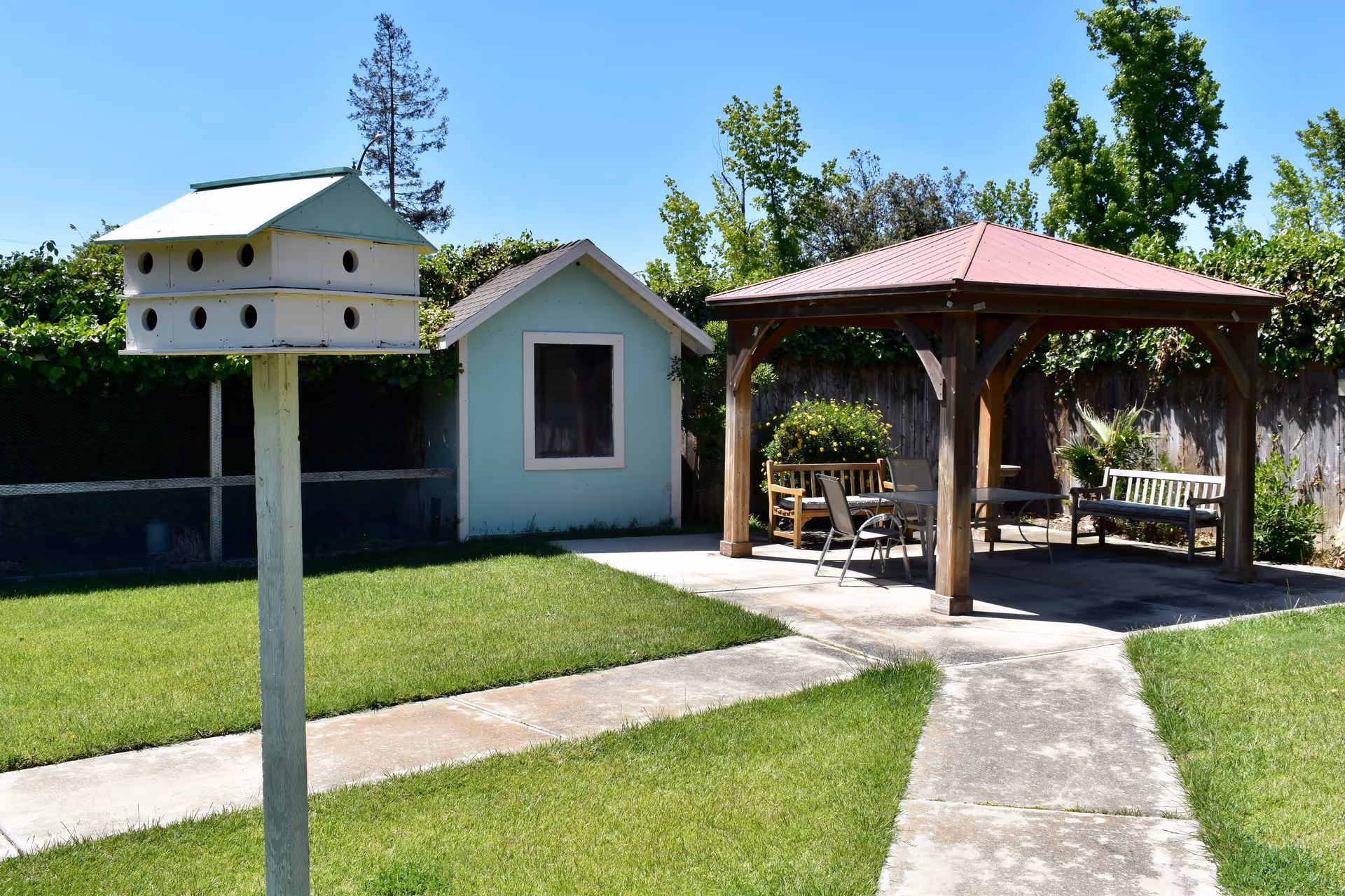 Outdoor garden area with a birdhouse on a post, a small light blue shed with a window, and a wooden gazebo with a red roof. Under the gazebo are benches and chairs arranged around a table. The area is surrounded by green grass, trees, and a wooden fence.