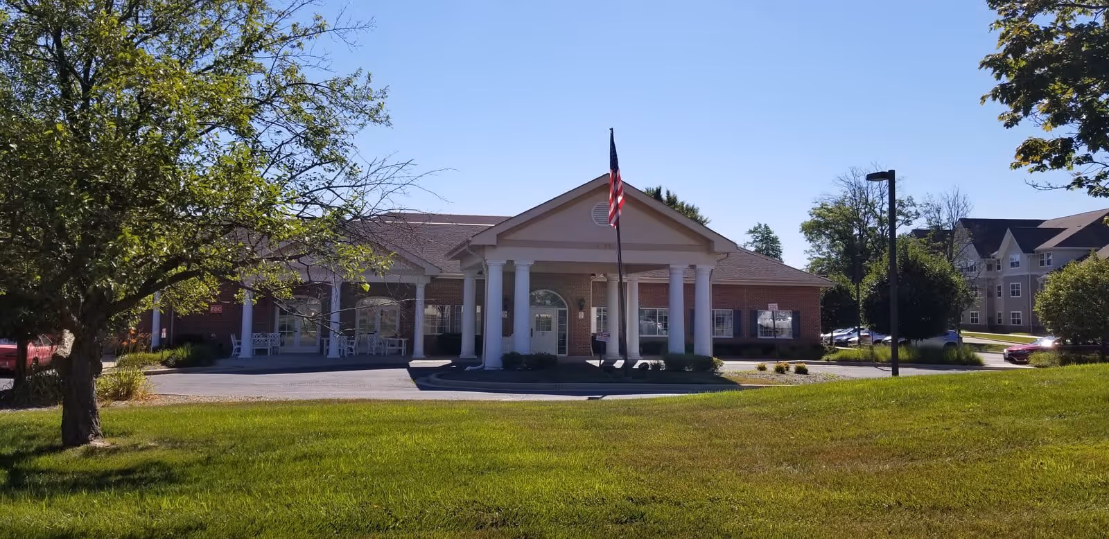Front exterior view of Meadow Park Rehabilitation And Healthcare Center building with a flagpole displaying the American flag in front. The building has white columns and a covered entrance, surrounded by green grass, trees, and a clear blue sky.