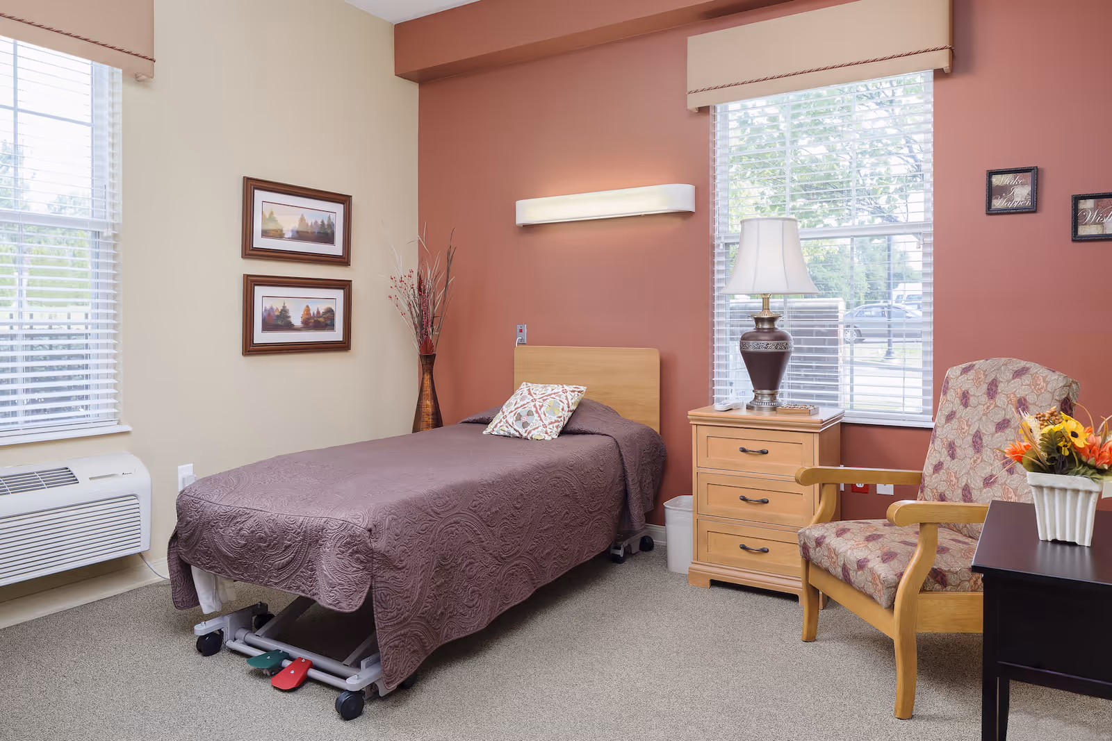 A senior living bedroom with a single bed covered in a purple quilt, a wooden nightstand with a lamp, a floral armchair, two windows with blinds, framed landscape pictures on the wall, and a small table with a flower arrangement.