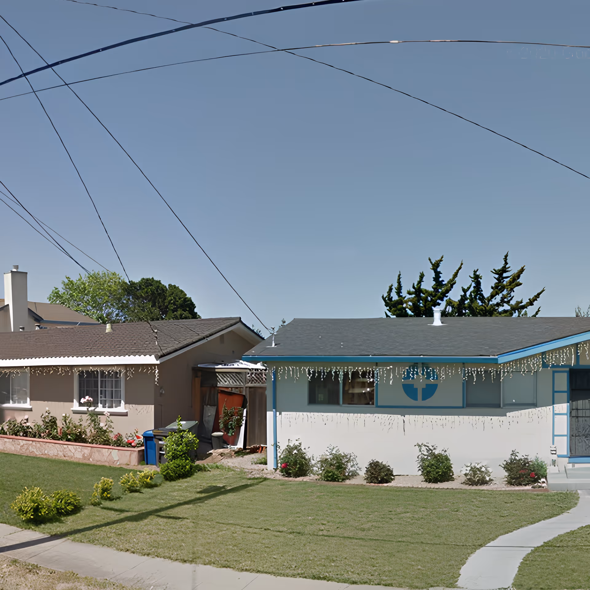 Exterior view of a single-story residential building with a well-maintained lawn and small bushes in front. The building has a gray roof with blue trim and a large window with a blue circular design. There are string lights hanging from the roof edge. Another house is partially visible to the left, and power lines cross the sky above.