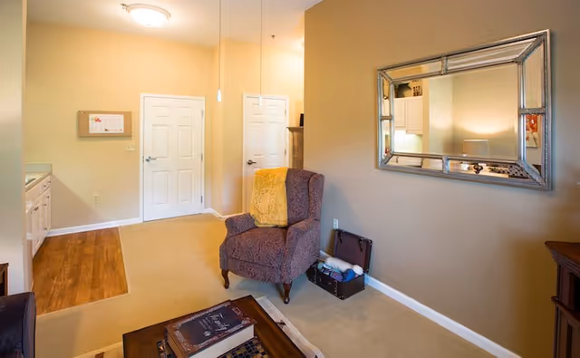 A cozy living room area with beige walls and carpet. There is a patterned armchair with a yellow throw blanket, a large decorative mirror on the wall, a wooden coffee table with books, and a small open chest with yarn and knitting supplies. Two white doors are visible in the background, along with a partial view of a kitchen area with wooden flooring.