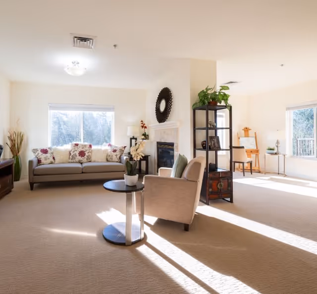 A bright and spacious living room with beige carpet and cream-colored walls. The room features a gray sofa with floral and white cushions, a beige armchair with a green pillow, a round side table with a potted orchid, a black shelving unit with decorative items and plants, and a fireplace with a circular mirror above it. Large windows allow natural light to fill the room.