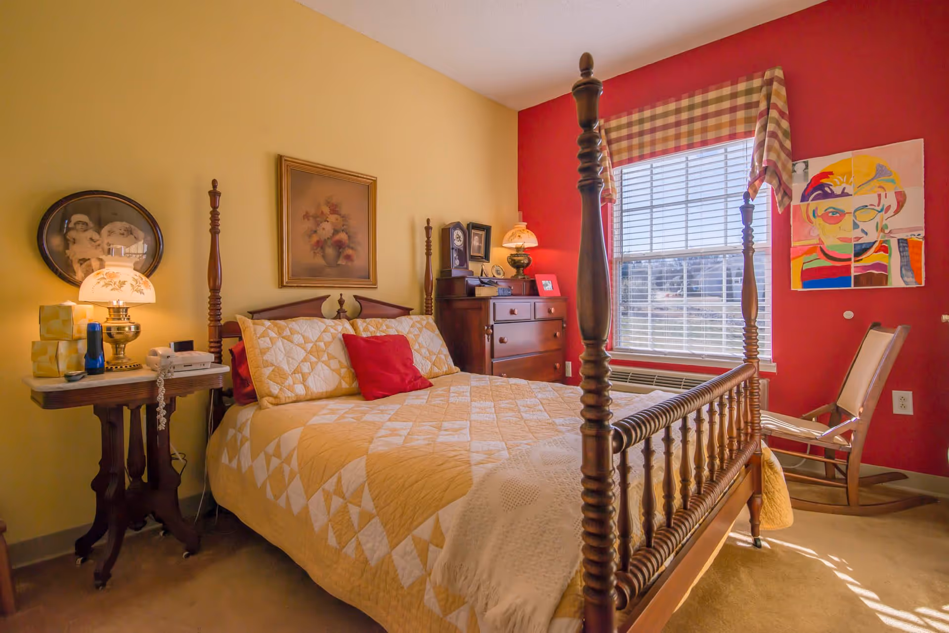 A cozy bedroom with a wooden four-poster bed covered in a yellow and white quilt, a red accent pillow, and a white throw blanket. The room has yellow and red walls, a window with plaid valance, a wooden dresser with a lamp and framed photos, a side table with a lamp, tissue box, and telephone, and a wooden rocking chair near the window. There are framed pictures on the walls, including a floral painting and a colorful portrait.