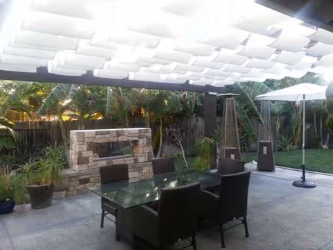 Under a white pergola, outdoor patio with glass table, six chairs, and stone fireplace.