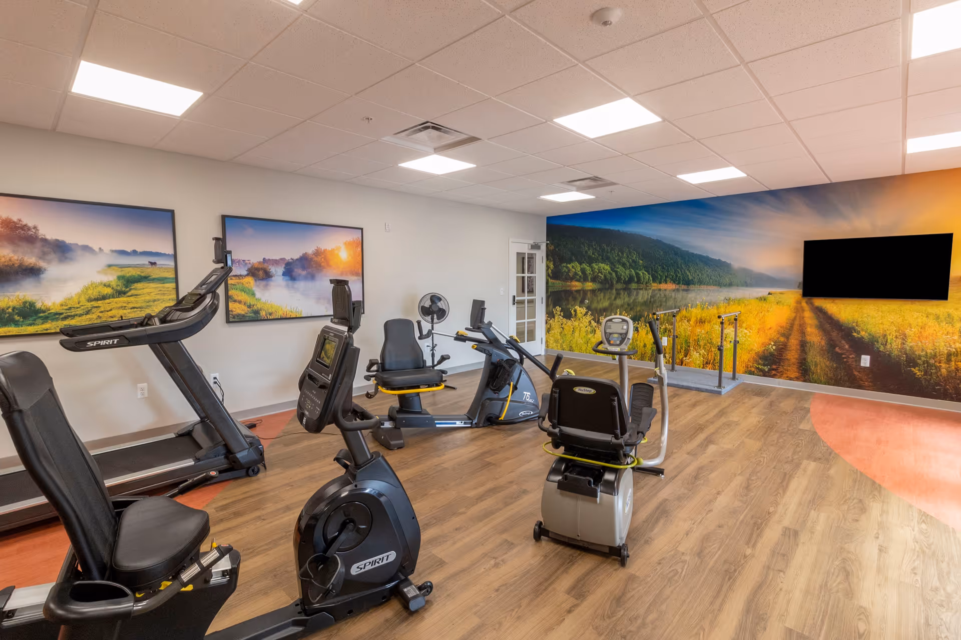 A fitness room with exercise equipment including recumbent bikes and a treadmill. The room has wood flooring and bright overhead lighting. The walls are decorated with large landscape photos and a mural of a scenic river and forest at sunrise. A flat screen TV is mounted on the mural wall.