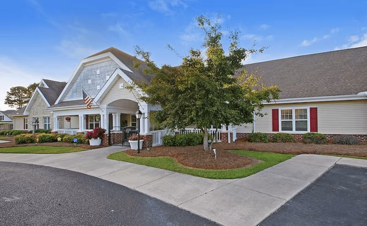 Exterior view of The Pines at Florence Assisted Living & Memory Care building with a well-maintained lawn, a tree, and a paved driveway under a clear blue sky.
