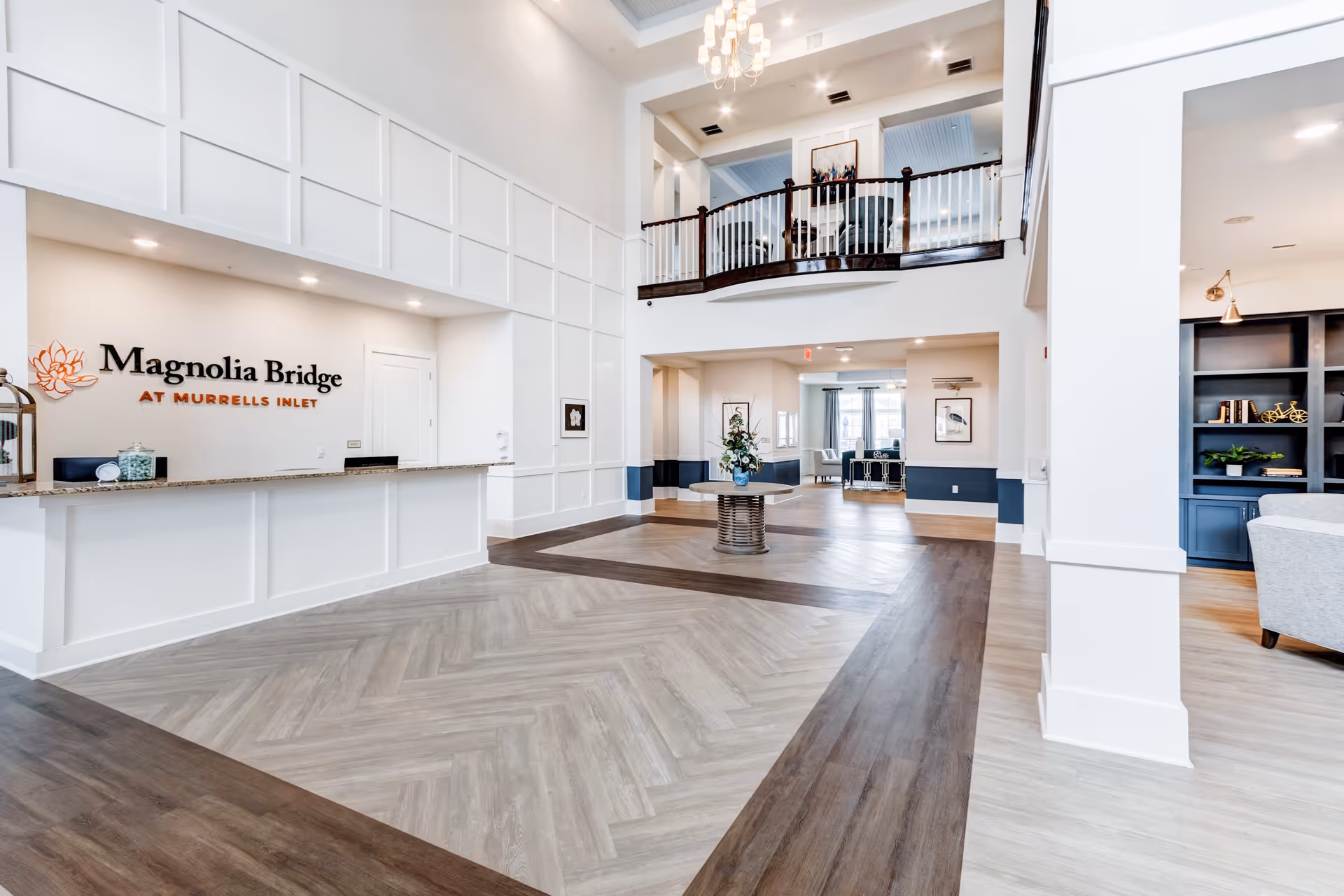 A spacious lobby area featuring a reception desk with the name 'Magnolia Bridge at Murrells Inlet' displayed. The floor has a unique herringbone pattern, and there is a decorative table with flowers in the center. In the background, seating areas are visible with large windows allowing natural light.