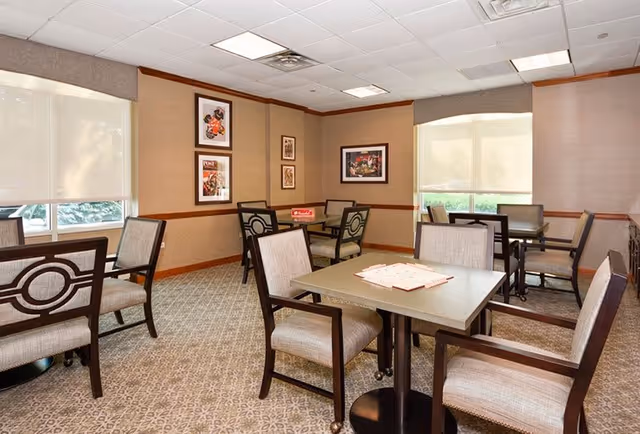 A well-lit dining area with several square tables and cushioned chairs arranged neatly. The room has beige walls with framed artwork, large windows with roller shades, and a patterned carpeted floor.