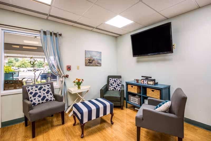 A small sitting area with three gray armchairs, each with a decorative pillow, arranged around a striped blue and white ottoman. A small white side table with a vase of flowers is next to one chair. A blue console table with wicker baskets and decorative items is against the wall beneath a mounted flat-screen TV. A window with blue and white curtains shows an outdoor view with chairs and greenery.