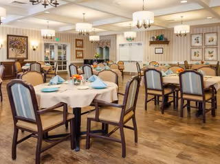Spacious, well-lit dining room with round tables set with plates, folded napkins, floral centerpieces, and wooden chairs.