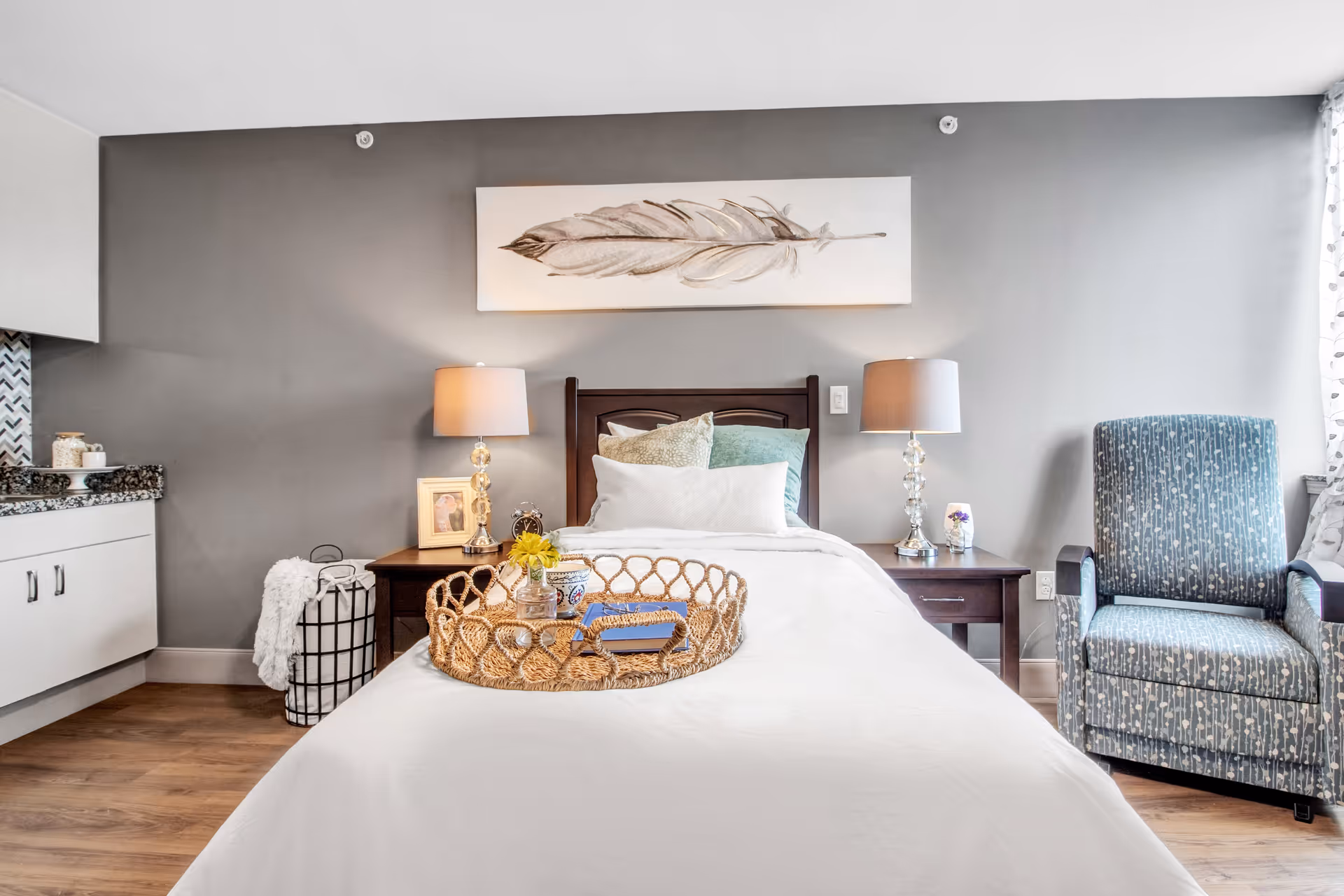 A cozy bedroom with a single bed covered in white bedding and decorative pillows. On the bed is a woven tray holding a small vase with yellow flowers and a book. There are two matching bedside tables with lamps on each side of the bed. A framed picture and an alarm clock are on the left bedside table. To the right of the bed is a blue patterned armchair. The walls are painted gray, and a large artwork of a feather hangs above the bed. Part of a kitchenette with white cabinets and granite countertop is visible on the left side of the image.