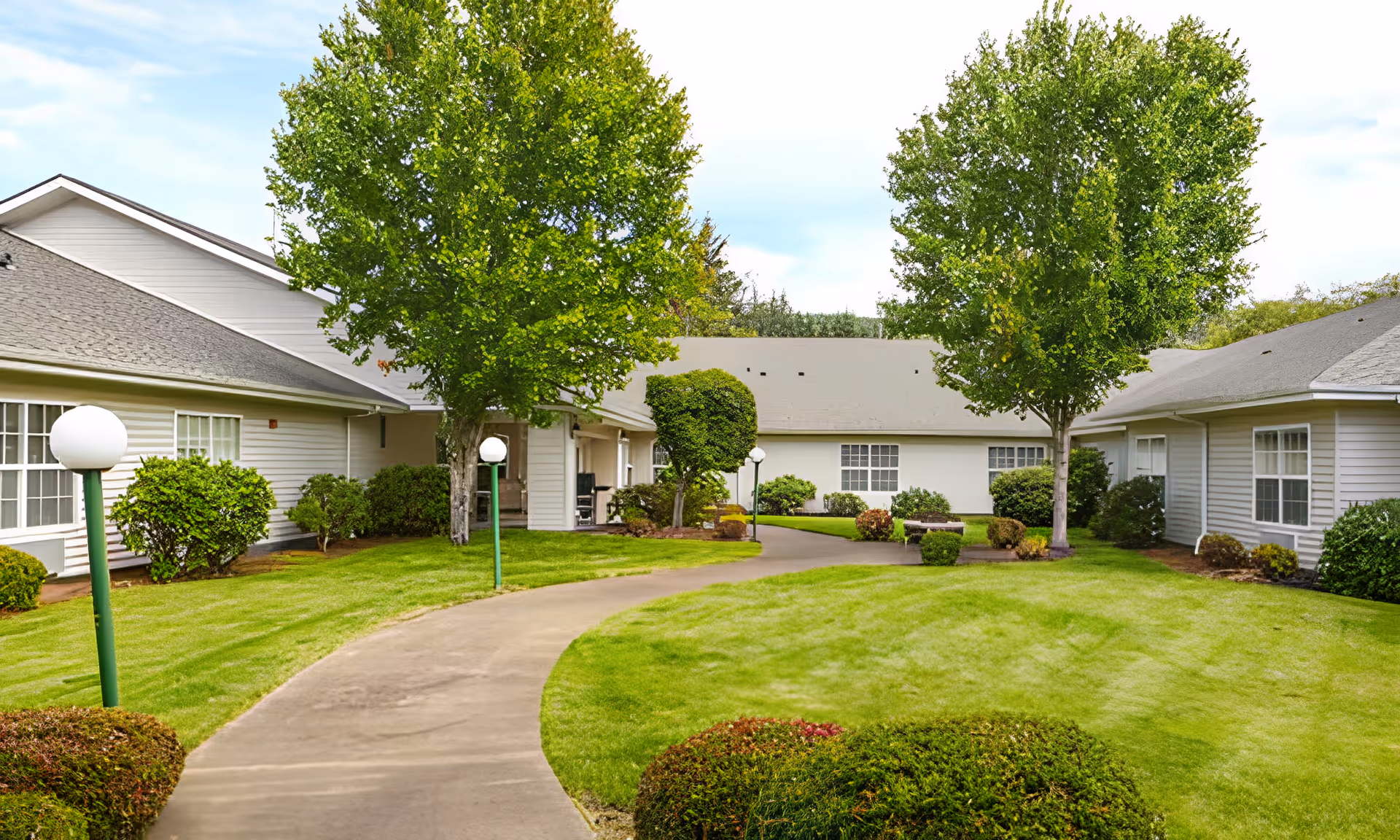 Courtyard of a single-story memory care facility with a curved paved walkway, manicured lawn, shrubs, trees, and lamp posts.