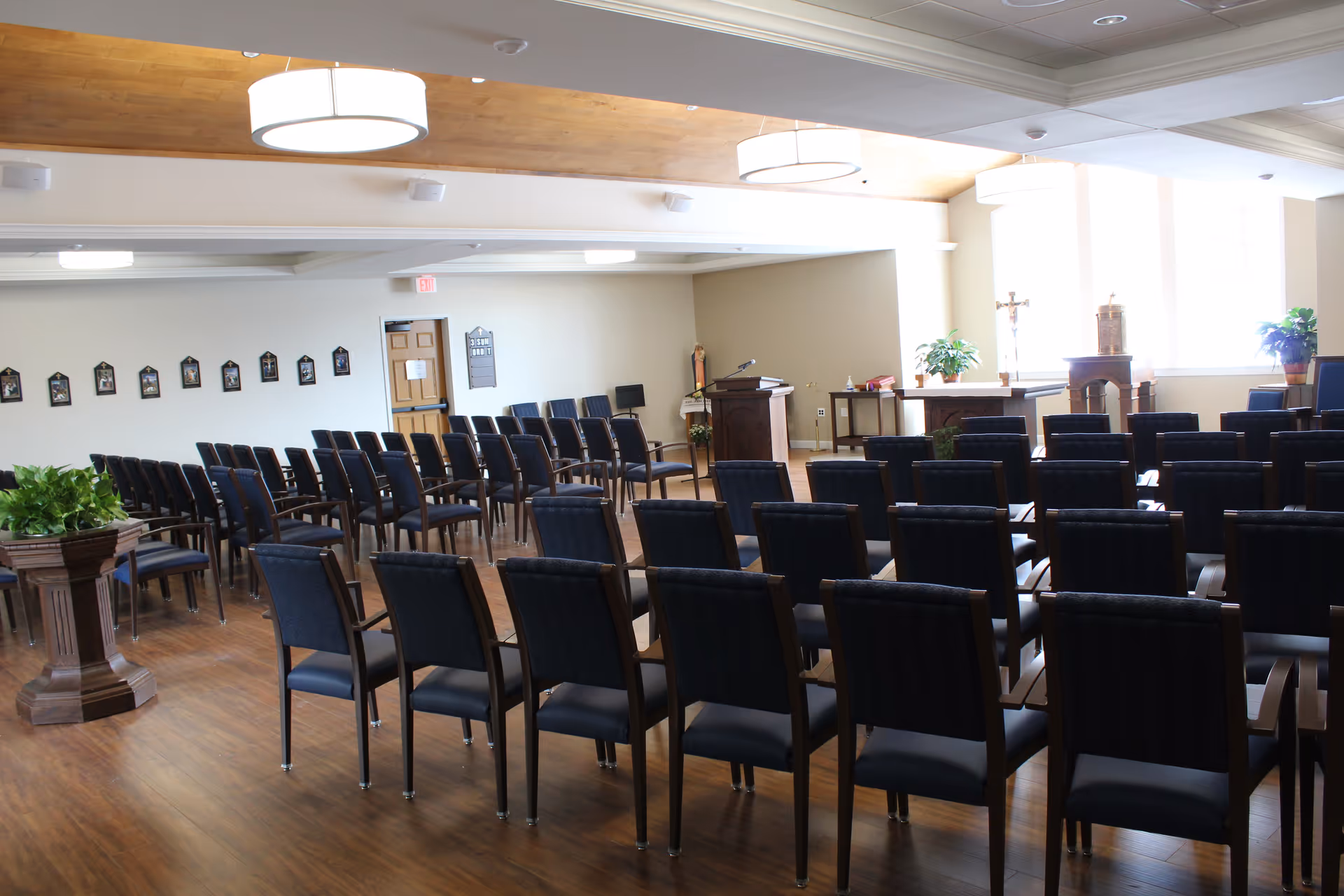 A chapel or meeting room with rows of black chairs arranged facing a wooden podium and altar area. The room has wooden flooring, light-colored walls, and large windows letting in natural light. Religious items such as a cross and statues are visible near the front.