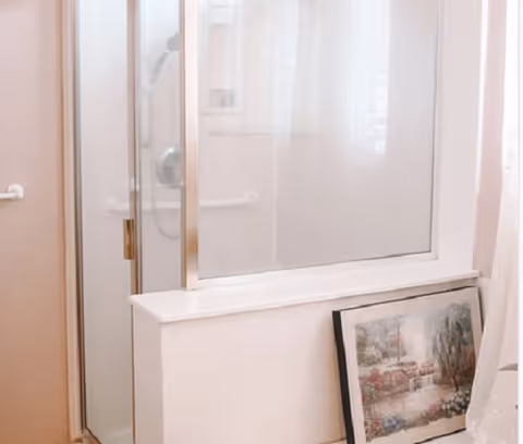 A bathroom corner featuring a glass shower enclosure with a metal frame, a white wall with a grab bar, and a framed landscape painting resting against the wall near a window with sheer curtains.