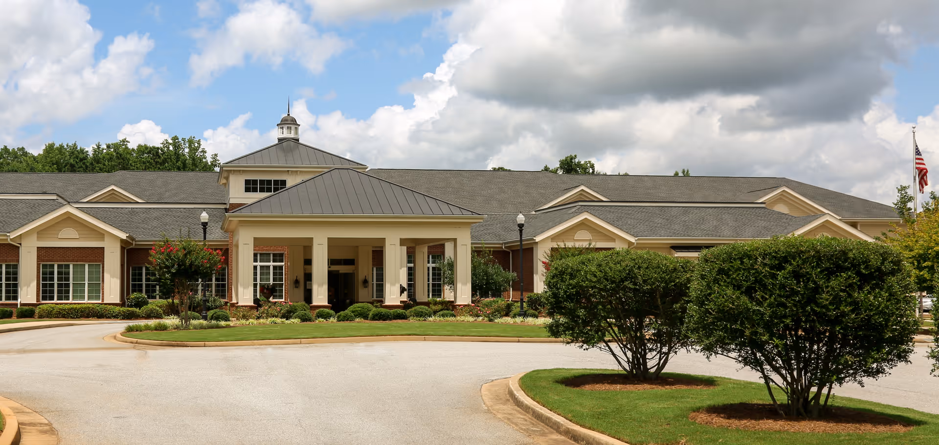 Front exterior of a single-story senior living facility with a covered portico entrance, circular driveway, and landscaped shrubs under a partly cloudy sky.