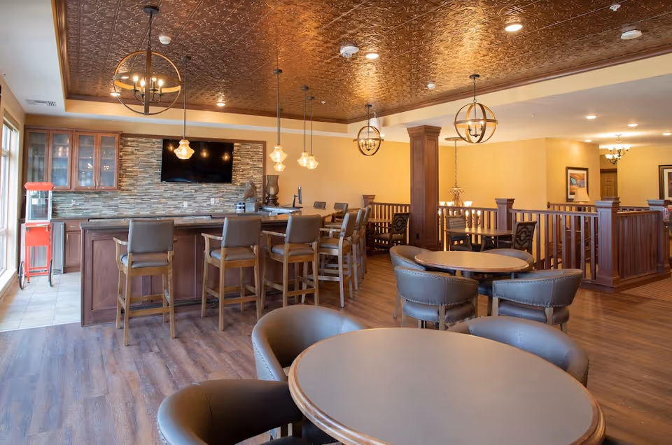 A spacious senior living common area with round tables and cushioned chairs in the foreground. In the background, there is a bar with high chairs, pendant lights hanging from a decorative ceiling, a mounted flat-screen TV, and a popcorn machine near large windows.