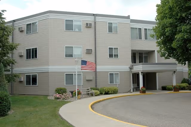 Exterior view of a three-story beige and white apartment building with multiple windows, an American flag on a flagpole near the entrance, a curved driveway, and some landscaping with bushes and trees.