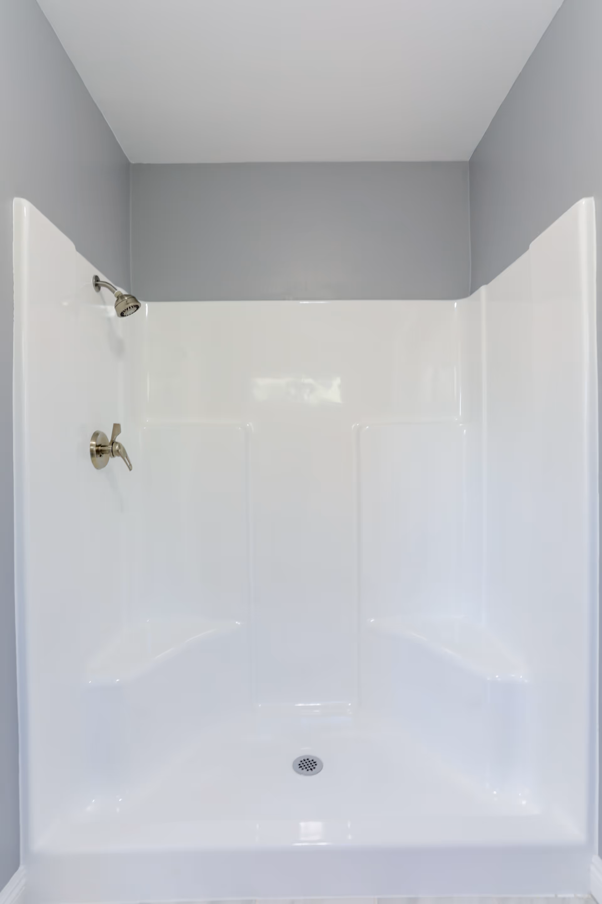 A clean, white shower stall with built-in corner seats on both sides, a silver showerhead, and a single handle control against light gray walls.