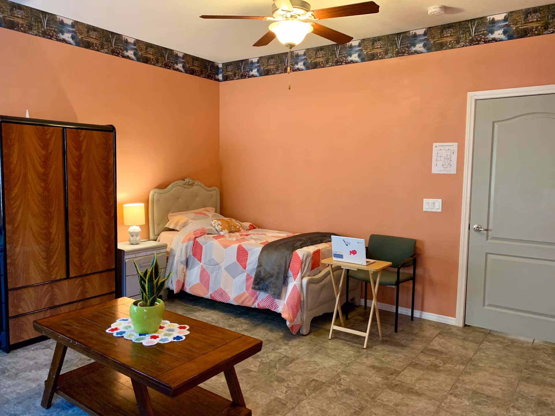 A cozy bedroom with a single bed covered in a geometric patterned quilt, a wooden wardrobe, a small nightstand with a lamp, a green chair, and a wooden coffee table with a potted plant. The walls are painted peach with a decorative border near the ceiling, and there is a ceiling fan with a light fixture.