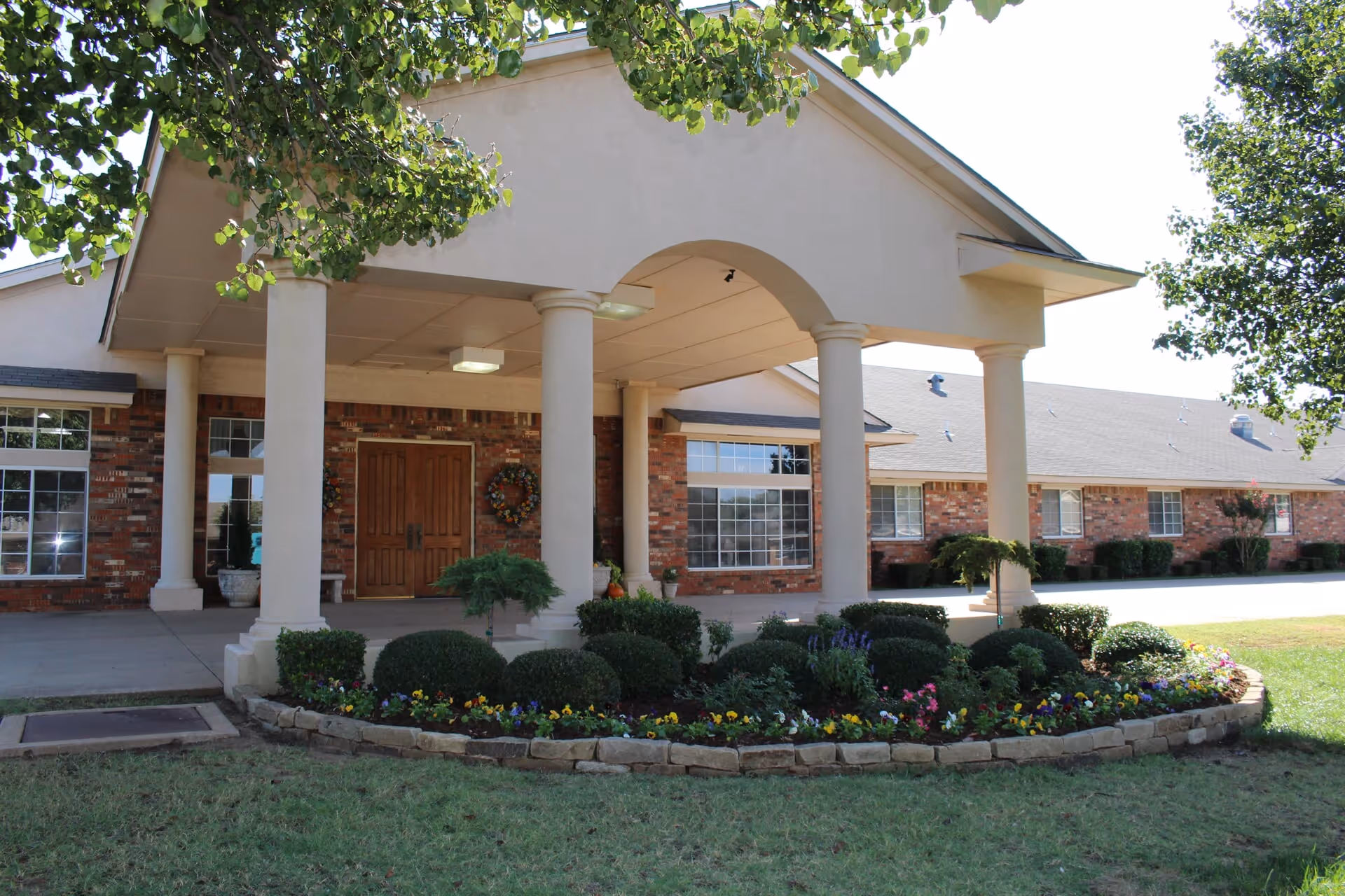 Front exterior view of a residential building with a covered entrance supported by columns, brick walls, large windows, and a landscaped flower bed with bushes and colorful flowers in front.