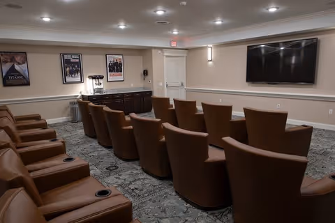 A small theater room with two rows of brown leather recliner chairs facing a large flat-screen TV mounted on the wall. The room has a carpeted floor, beige walls, and ceiling lights. There is a counter with a popcorn machine and framed movie posters on the wall behind it.