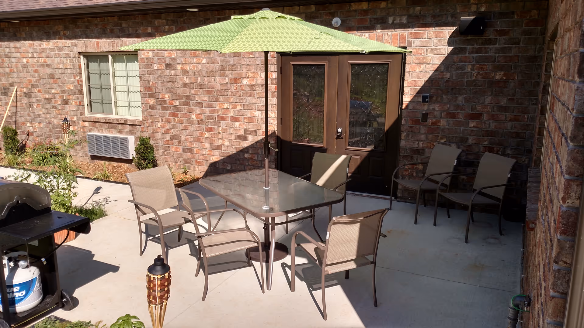 Outdoor patio area with a glass-top table surrounded by six beige chairs. A large green umbrella provides shade over the table. There is a barbecue grill on the left side and a brick building with double glass doors and a window in the background.