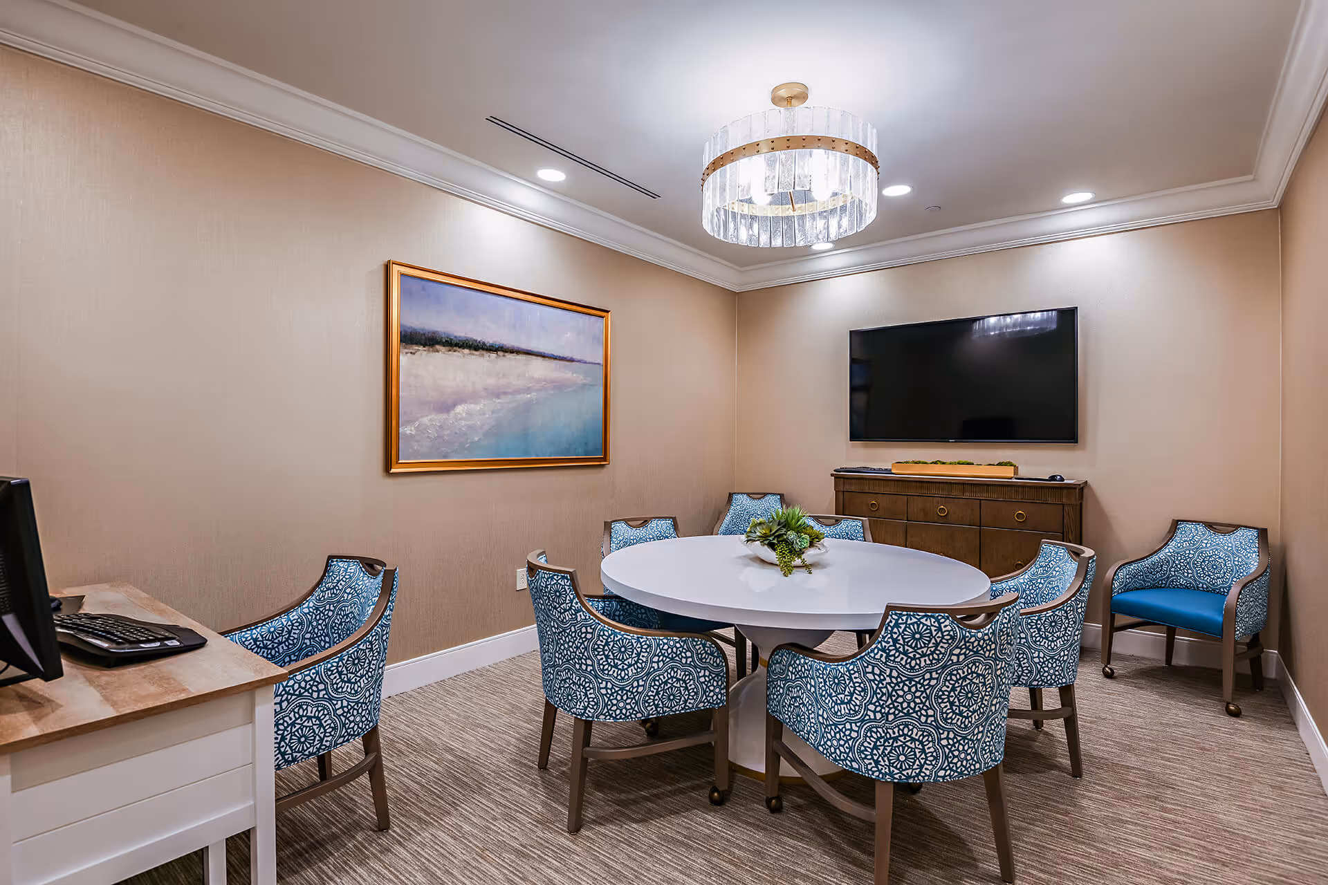 Small meeting/dining room with a round white table surrounded by patterned blue chairs, a wall-mounted TV, sideboard, and framed artwork.
