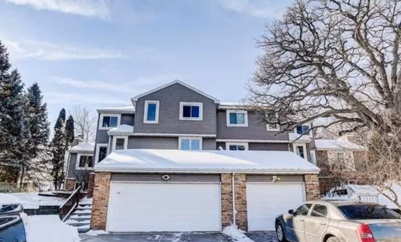 Snow-covered multi-unit residential building with garages and a parked car.
