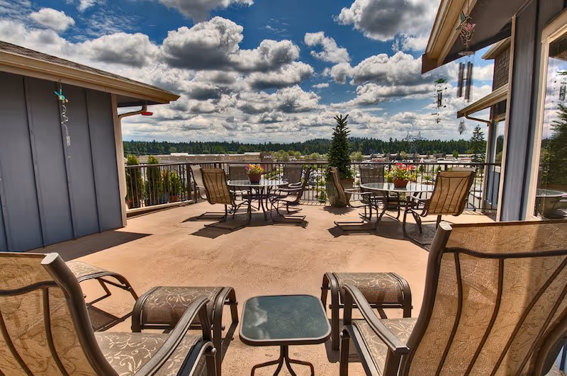 Outdoor patio area with several metal tables and chairs, some with potted plants on top. The patio overlooks a scenic view of trees and a partly cloudy sky. There are wind chimes hanging from the roof edges.