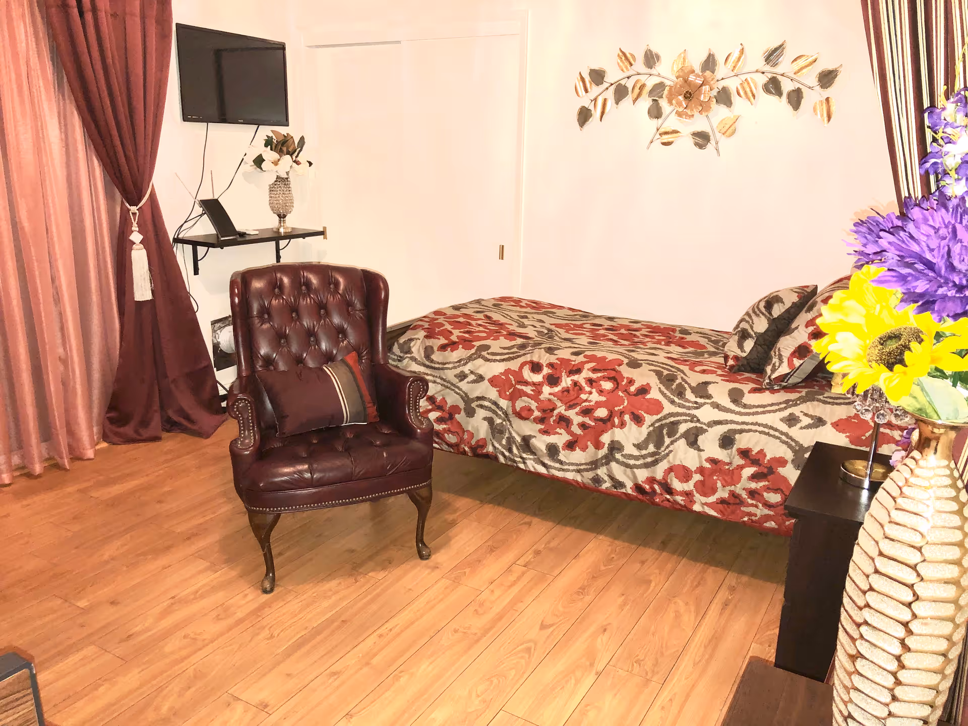 Cozy bedroom with a single bed covered in a patterned bedspread, a tufted leather armchair, wall-mounted TV, and flowers on a side table.