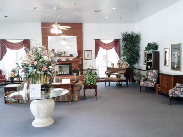 Spacious assisted-living common room with sofas, a fireplace, a piano, and a large floral centerpiece on a glass table.