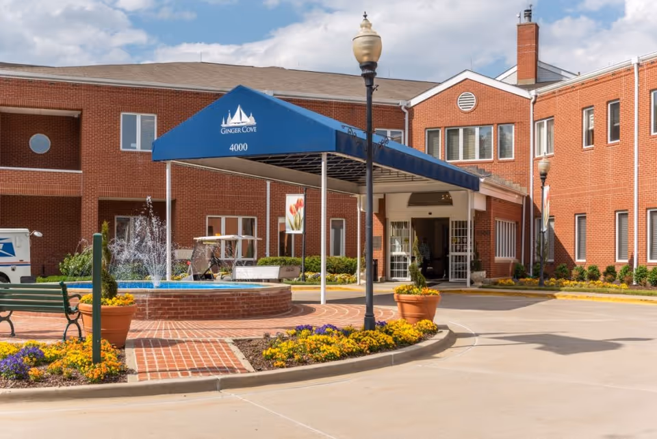 Entrance of Ginger Cove facility featuring a blue canopy with the facility name and number 4000, a brick building, a water fountain surrounded by flowers, benches, and a street lamp.