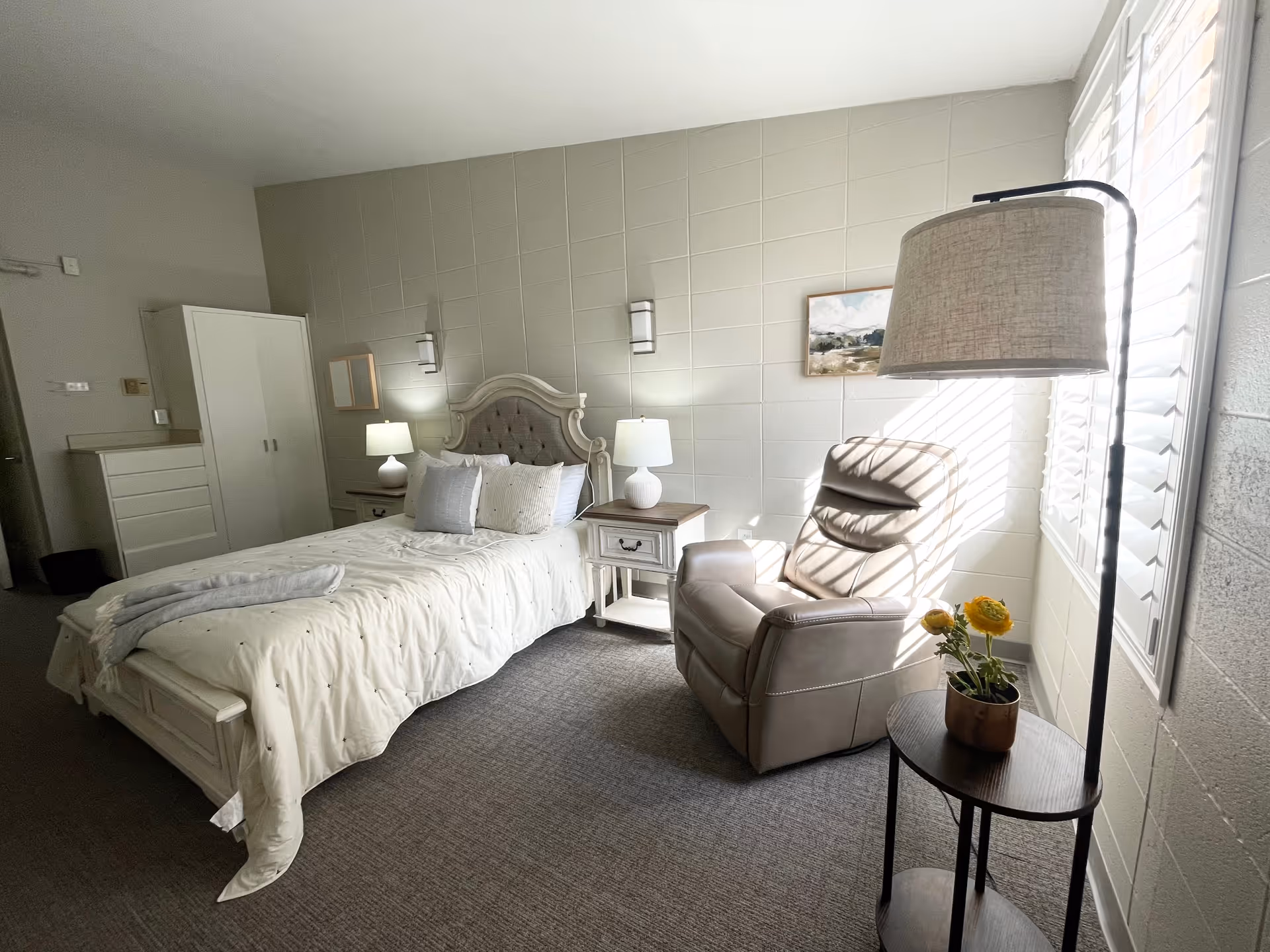 A bright, cozy bedroom with a neatly made bed featuring white bedding and multiple pillows. On either side of the bed are nightstands with lamps. To the right, there is a comfortable beige recliner chair next to a small round table with a potted yellow flower and a tall floor lamp. The room has light-colored walls with a framed picture and a window with white shutters letting in natural light.