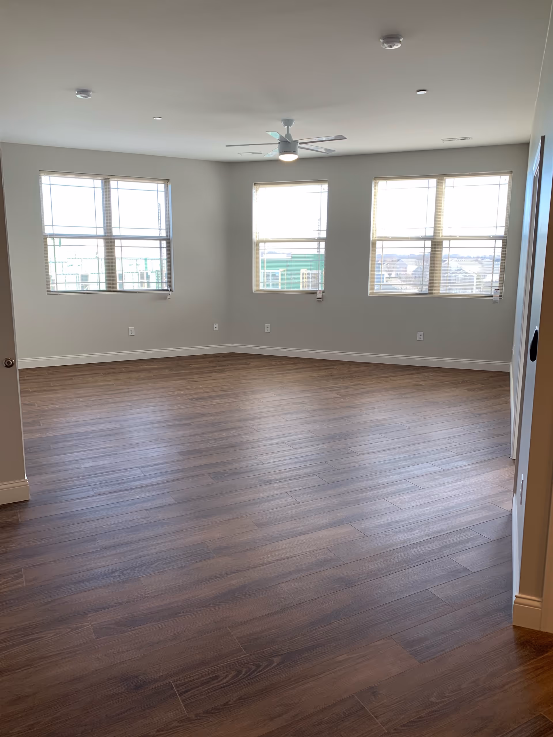 Empty room with wood flooring, three large windows with blinds, white walls, a ceiling fan with light, and multiple electrical outlets along the walls.