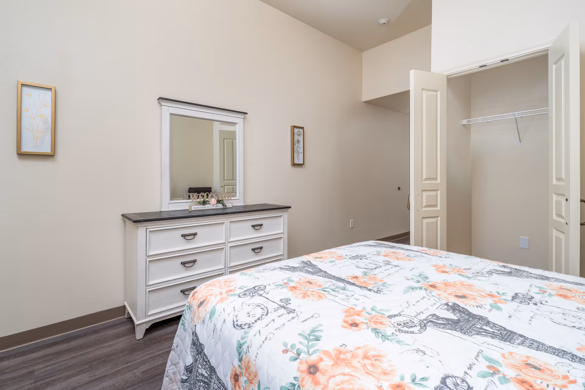 A bedroom with a floral patterned bedspread in the foreground, a white dresser with a mirror on the wall above it, two framed floral artworks on the wall, and an open closet with bi-fold doors.
