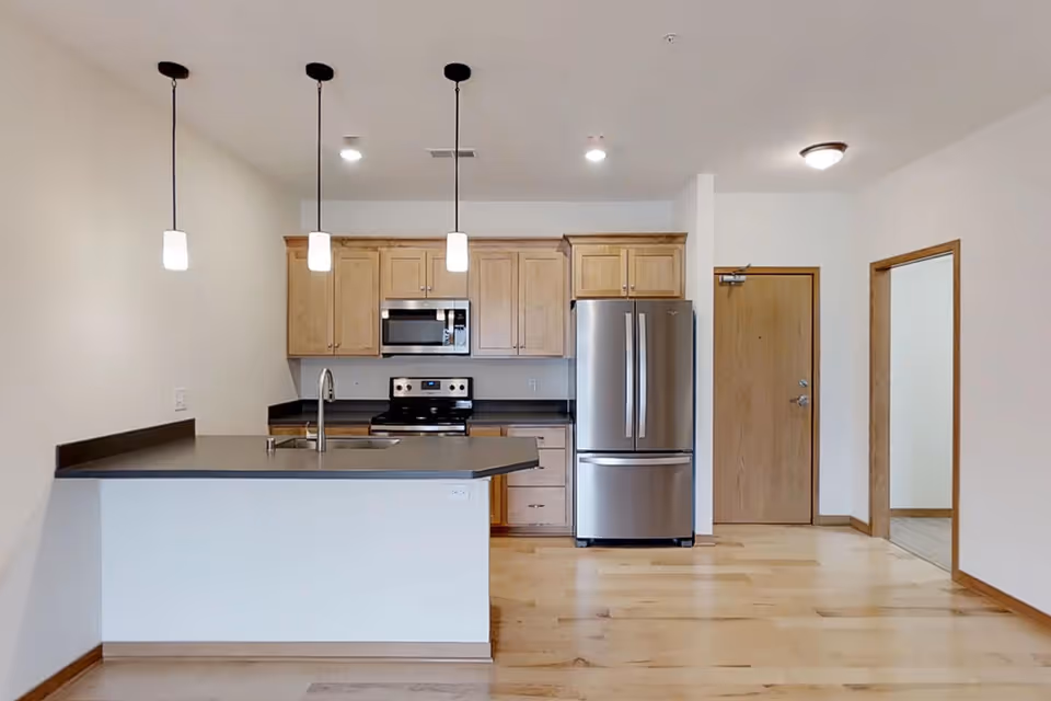 Modern kitchen area with light wood cabinets, stainless steel refrigerator, stove, and microwave. A kitchen island with a sink and three pendant lights hanging above. Light wood flooring and white walls with a wooden door and an open doorway visible.