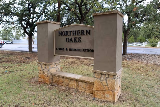 Stone and stucco sign for Northern Oaks Living & Rehabilitation Center situated on a grassy area with trees and a parking lot in the background.