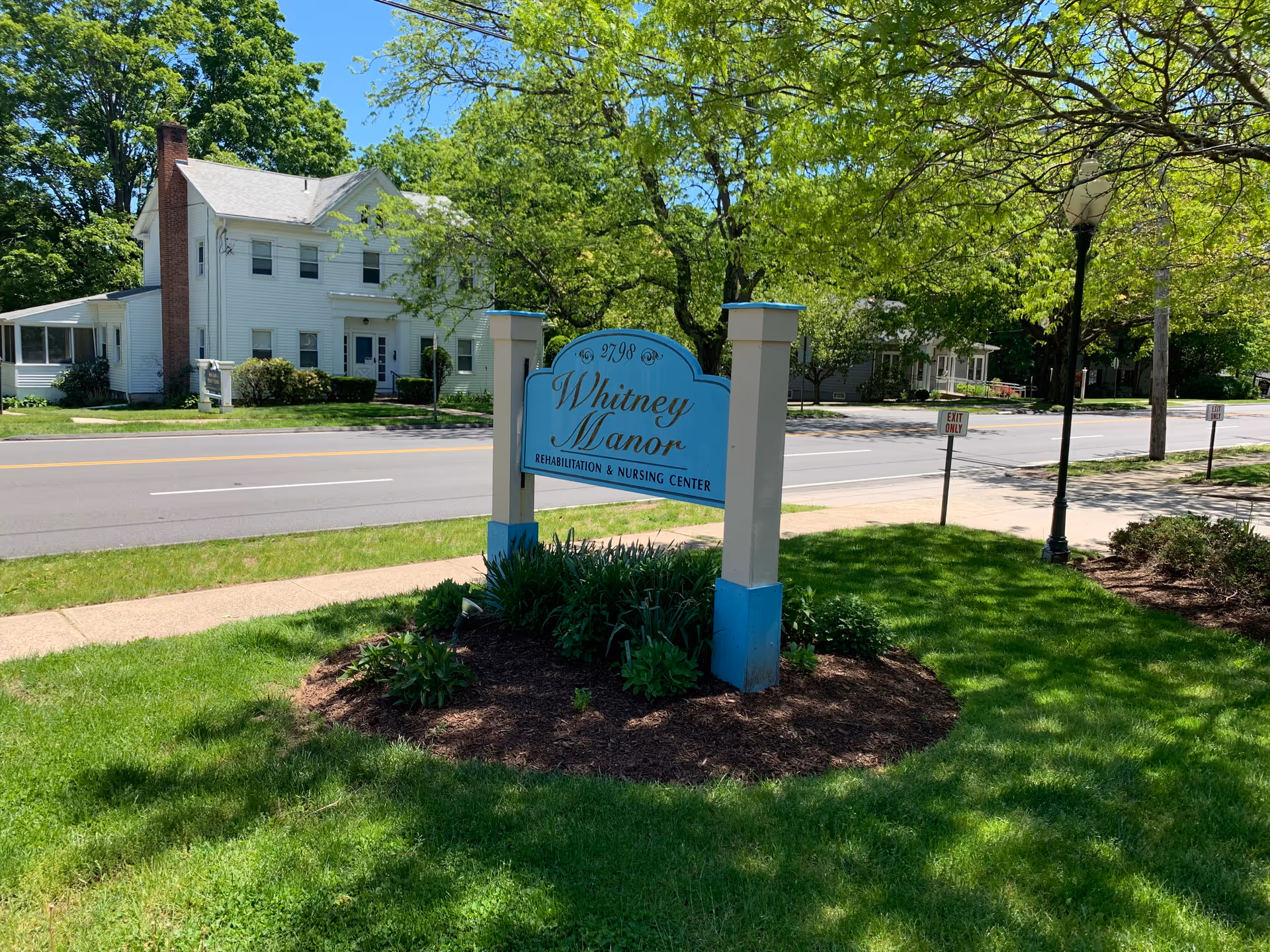 A blue Whitney Manor rehabilitation and nursing center sign on a grassy front lawn with a street and houses in the background.