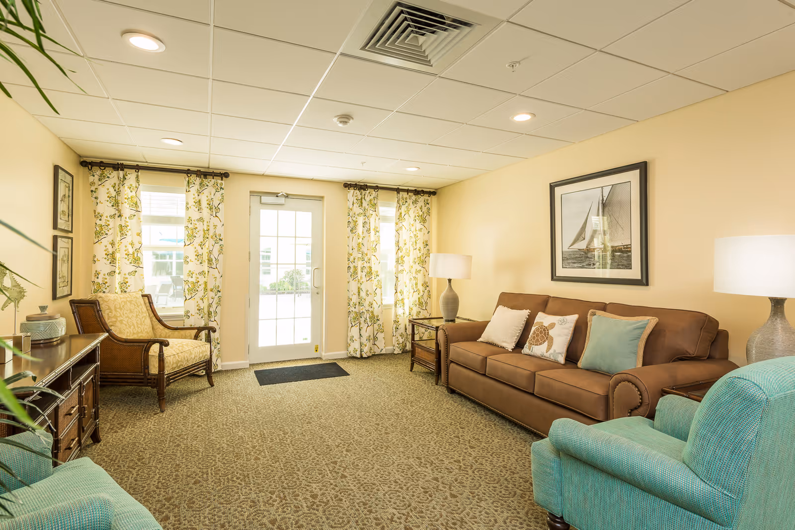Bright cozy living room with a brown sofa, teal armchairs, patterned curtains, lamps, and a glass door to the outside.