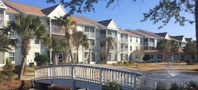 Exterior view of a multi-story senior living facility with balconies, palm trees, a small pond with a fountain, and a white wooden footbridge in the foreground under a clear blue sky.