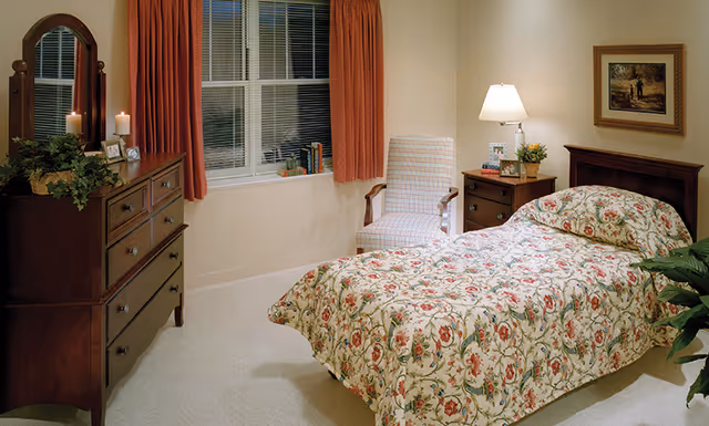 A tidy, traditionally furnished bedroom with a single floral bed, wooden dresser and nightstand, armchair, lamp, and a curtained window.