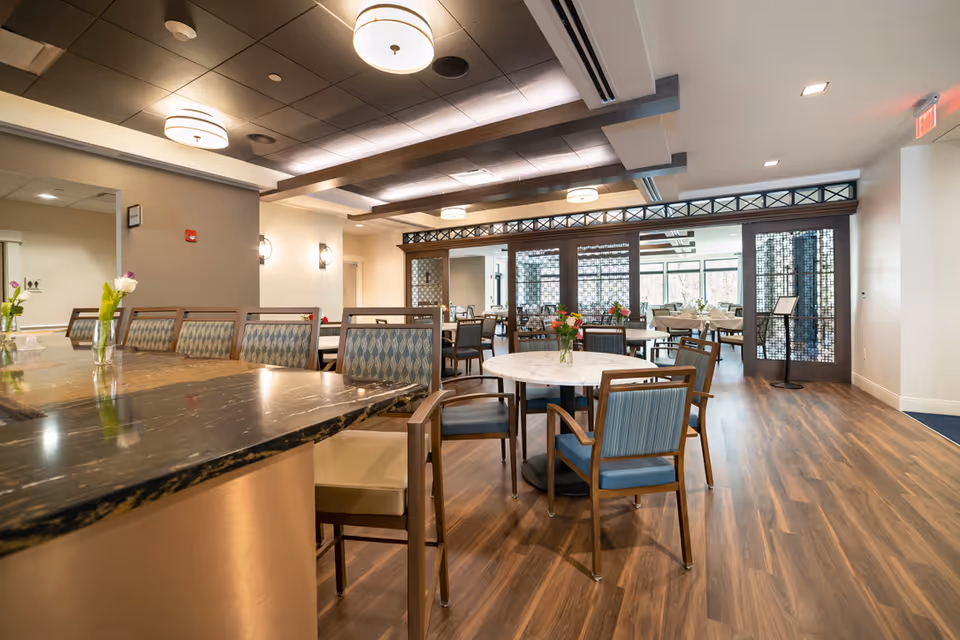 Interior view of a dining area in Briarcliffe Preserve Assisted and Independent Living featuring wooden floors, multiple tables with chairs, some with floral centerpieces, and modern ceiling lights. The space is well-lit with natural light coming through large windows in the background.