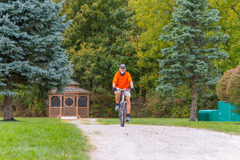 An elderly man wearing an orange shirt and cap rides a bicycle on a gravel path surrounded by green grass and trees. A wooden gazebo is visible in the background among the trees.