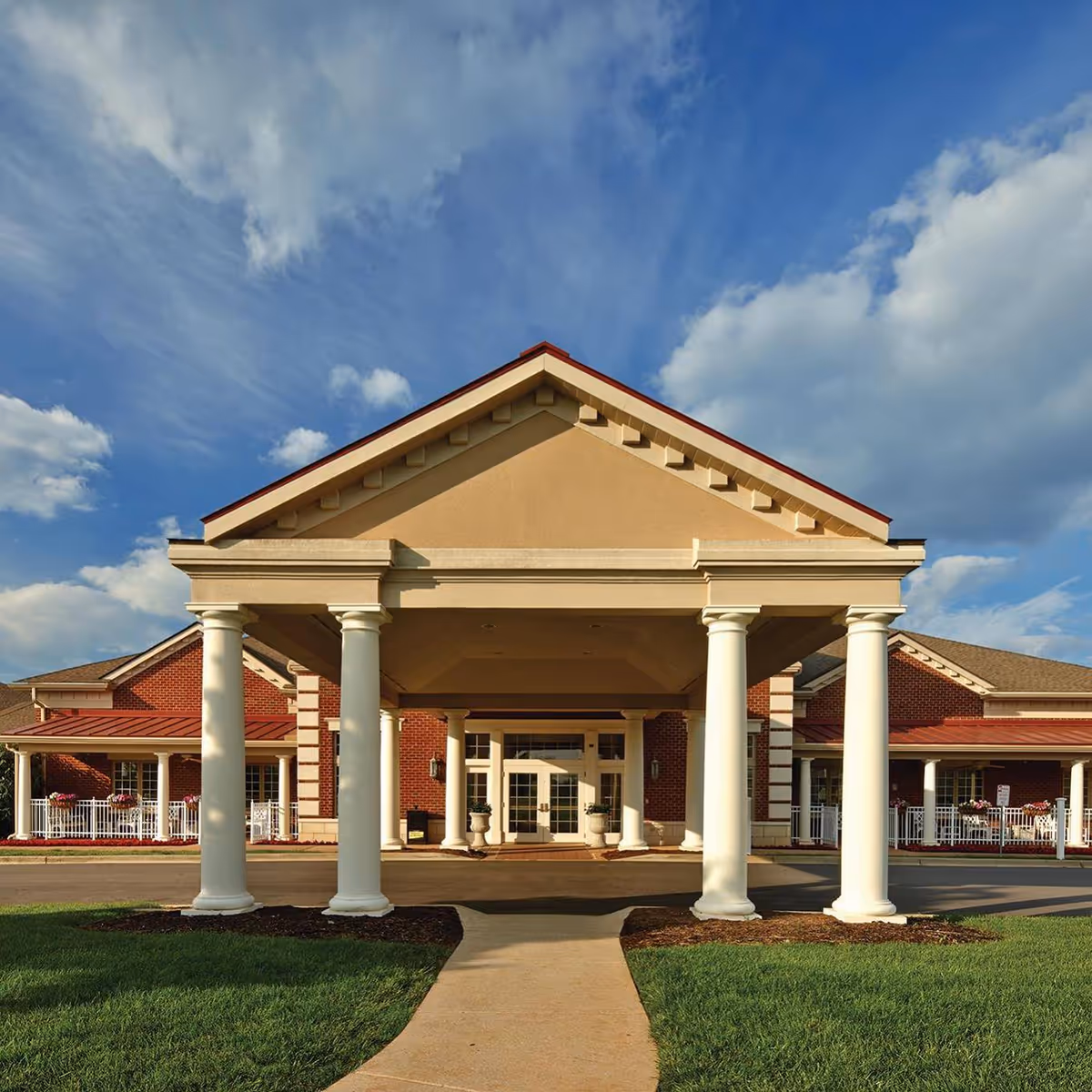 Front exterior view of Valhalla Post Acute facility featuring a covered entrance with large white columns, a brick building facade, and a clear blue sky with some clouds.