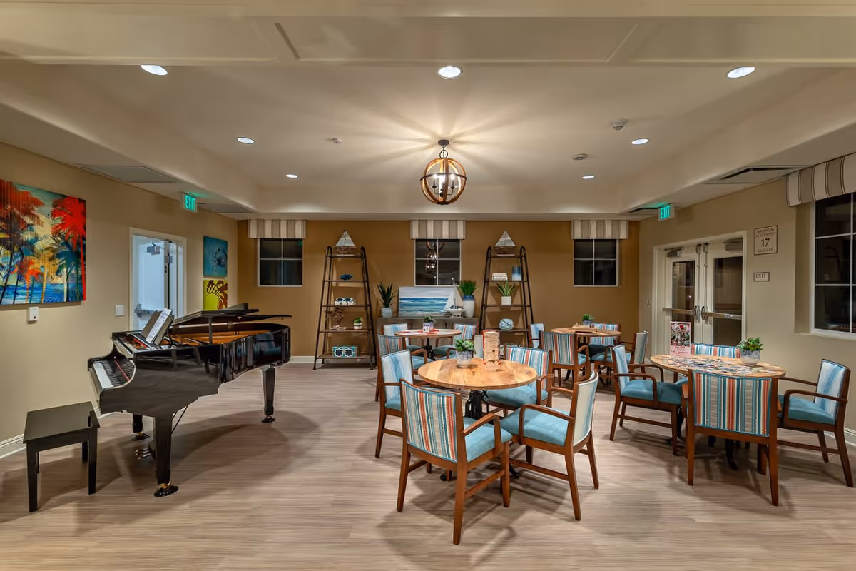A well-lit common area in a senior living facility featuring several round wooden tables with striped upholstered chairs. A black grand piano is positioned on the left side of the room. The walls are decorated with colorful paintings and shelves holding decorative items. There are windows and exit signs visible in the background.