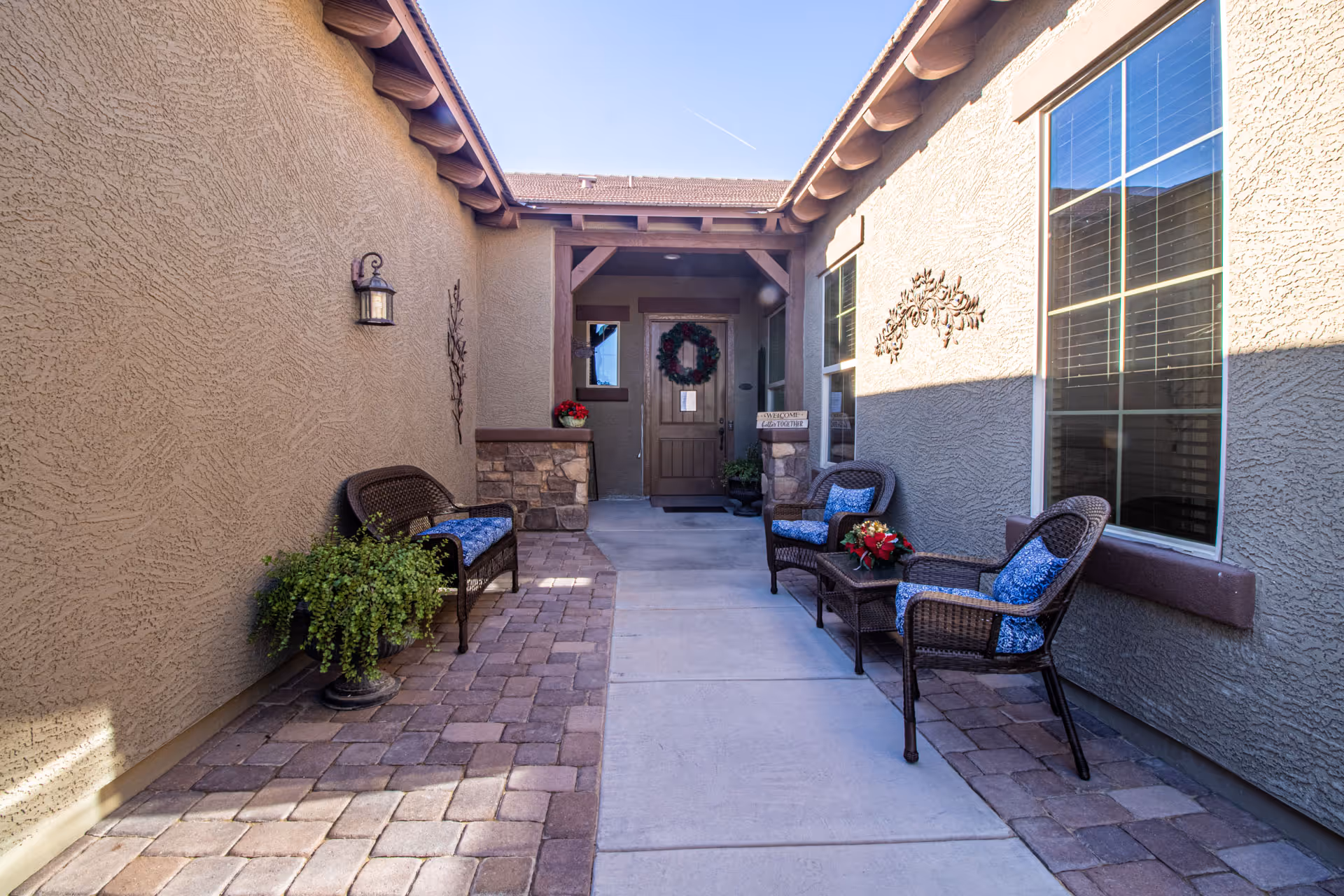 Outdoor entrance area of Pebble Heights Assisted Living & Memory Care featuring a pathway leading to a wooden door decorated with a wreath. The area is flanked by beige stucco walls with windows and outdoor wall lamps. There are wicker chairs with blue cushions and a small table with a flower arrangement on the right side, and a wicker bench with blue cushions and a potted plant on the left side.